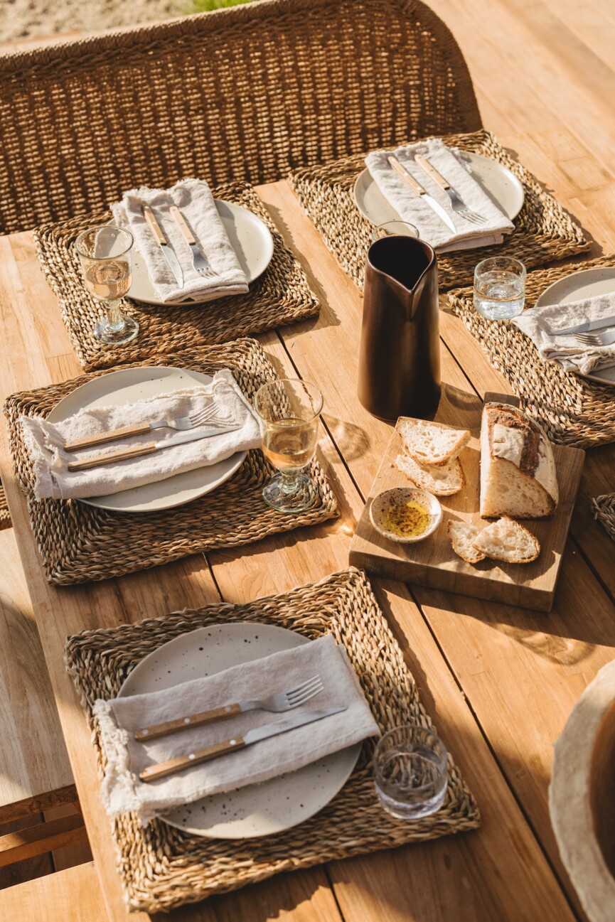 Sunlit wood dining table with woven placemats, white plates, linen napkins, and a dark pitcher.