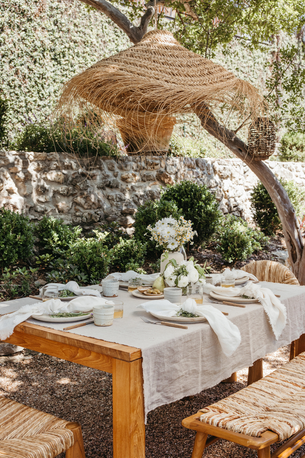 Wood outdoor dining table with wood bench and raffia pendant set with gray linen tablecloth neutral dinnerware and white napkins.