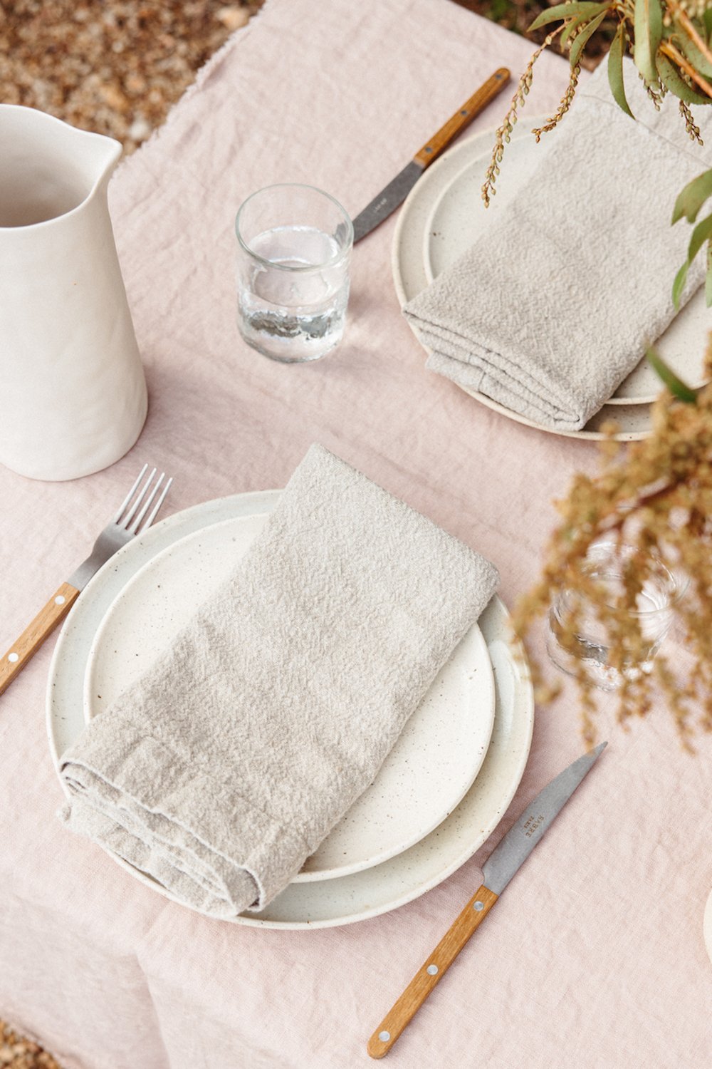 Simple table setting on pink linen tablecloth with two white plates, gray linen napkins, forks, and knives.