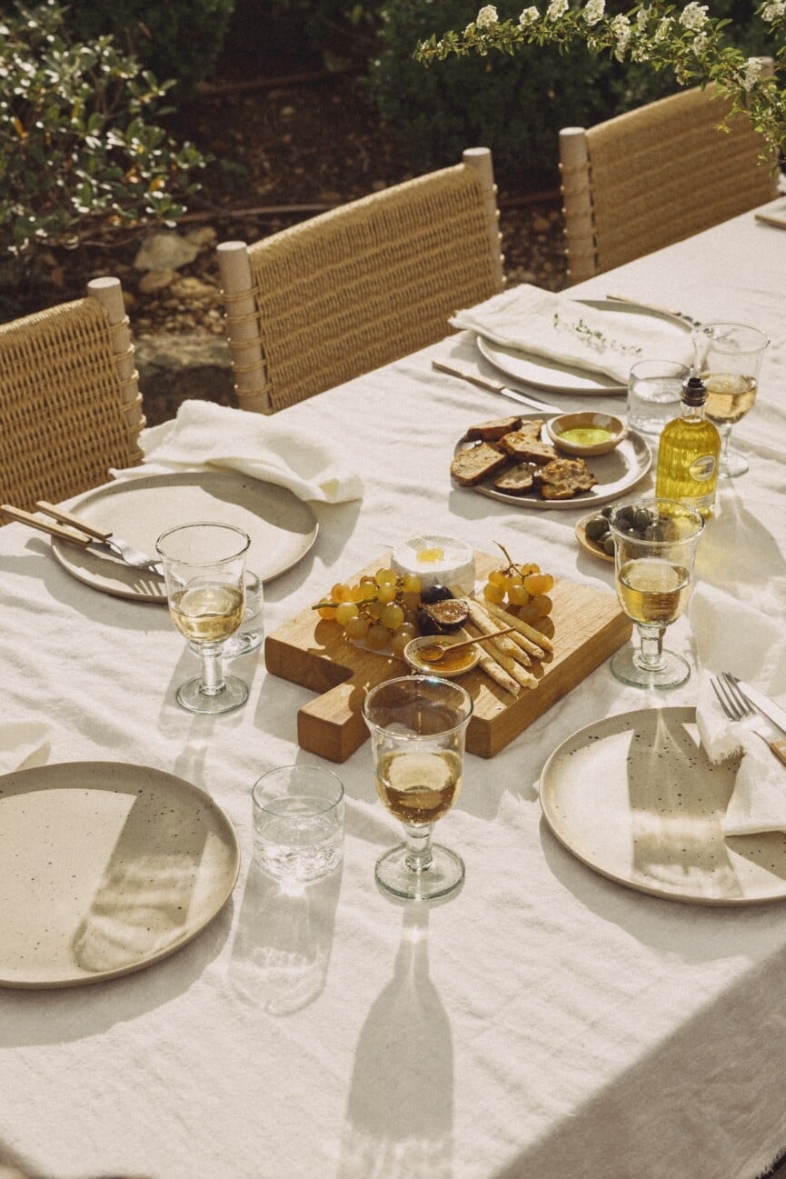 Garden lunch table with pale plates, clear goblets, and a wood serving board holding grapes and appetizers.