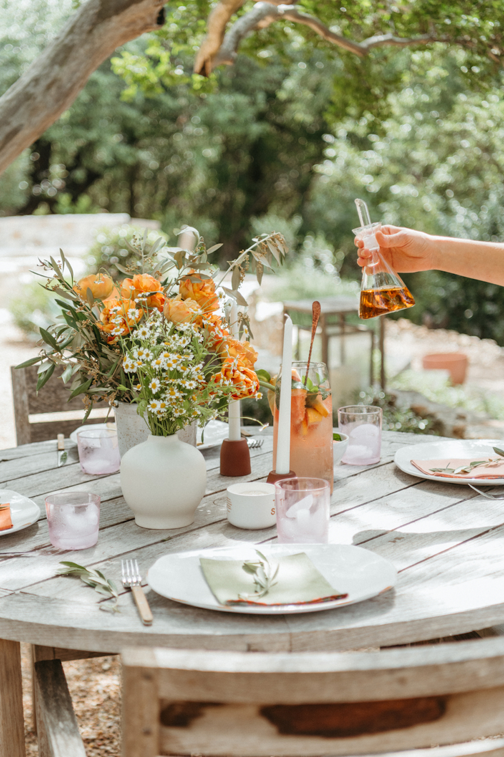 Sunny outdoor dining table with white plates, gray linen napkins, simple flatware, and an arrangement of eucalyptus, orange flowers, and daisies.