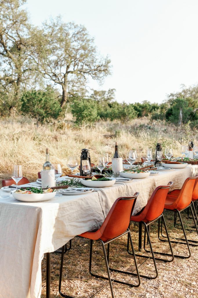 Una mesa larga al aire libre puesta en el campo con un mantel de lino neutro, sillas rojas, copas de vino y comida en platos blancos.