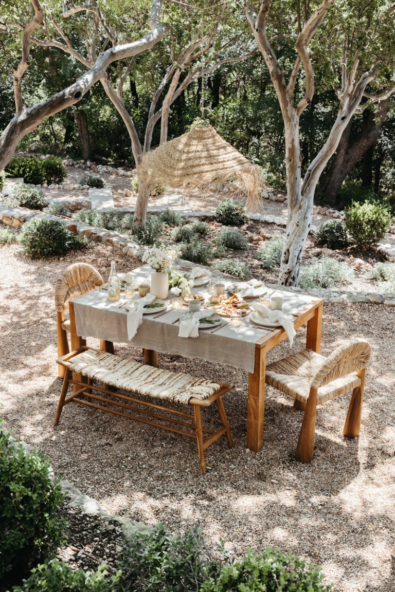 Mesa exterior natural en un patio trasero bañado por el sol con mantel de lino neutro, jarrón blanco con flores, platos neutros y servilletas blancas.