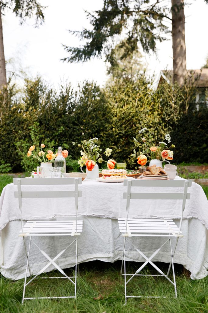 Pequeña mesa de comedor exterior cuadrada con mantel de lino blanco, varios platos y cristalería, y flores naranjas y blancas.