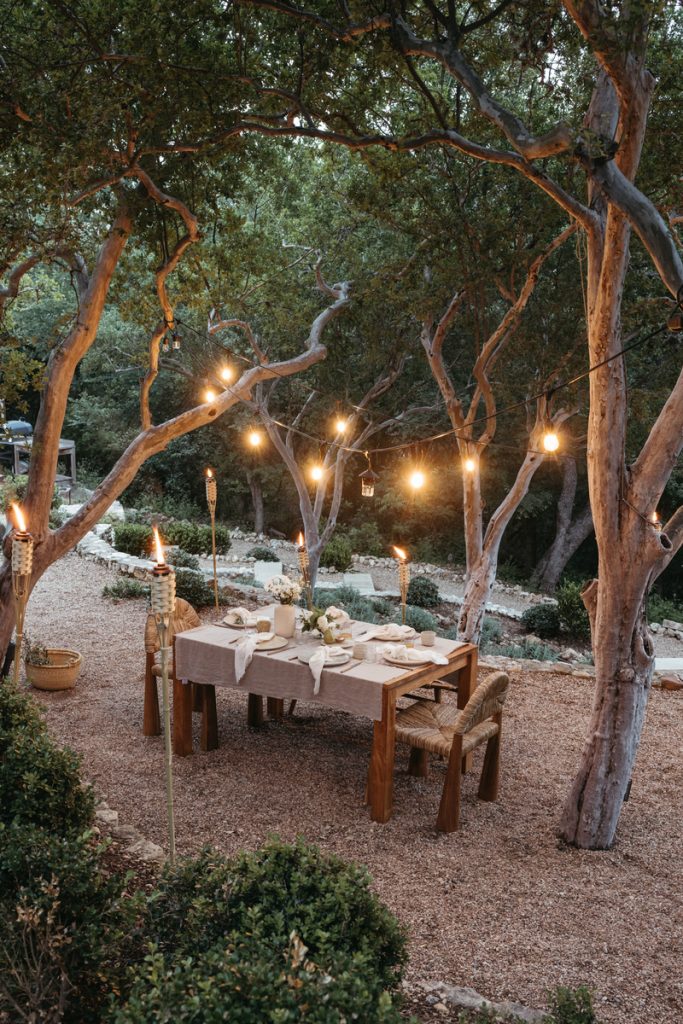 Outdoor table setting at night beneath string of cafe lights strung between trees and surrounded by tiki torches.