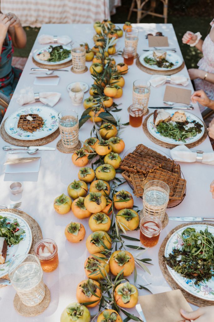 Juego de mesa de comedor rectangular con mantel rosa, platos tejidos, platos florales y un centro horizontal de caquis naranjas.