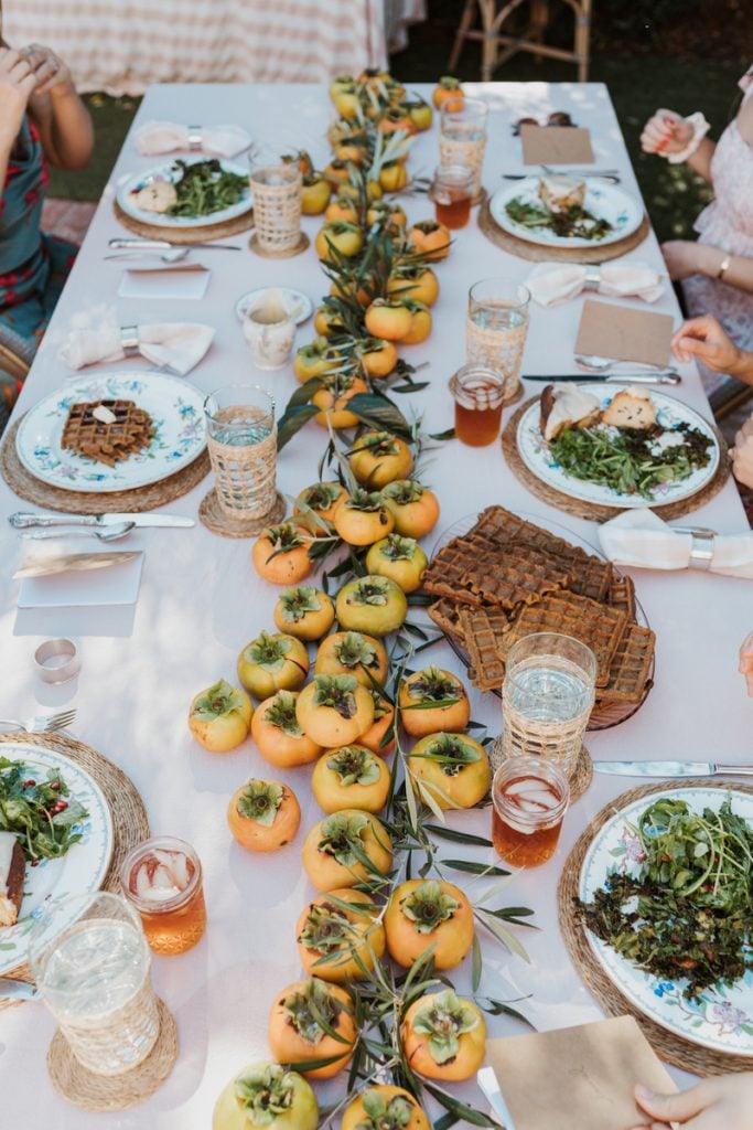 Rectangular dining table set with pink tablecloth, woven chargers, floral plates, and a horizontal centerpiece of orange persimmons.