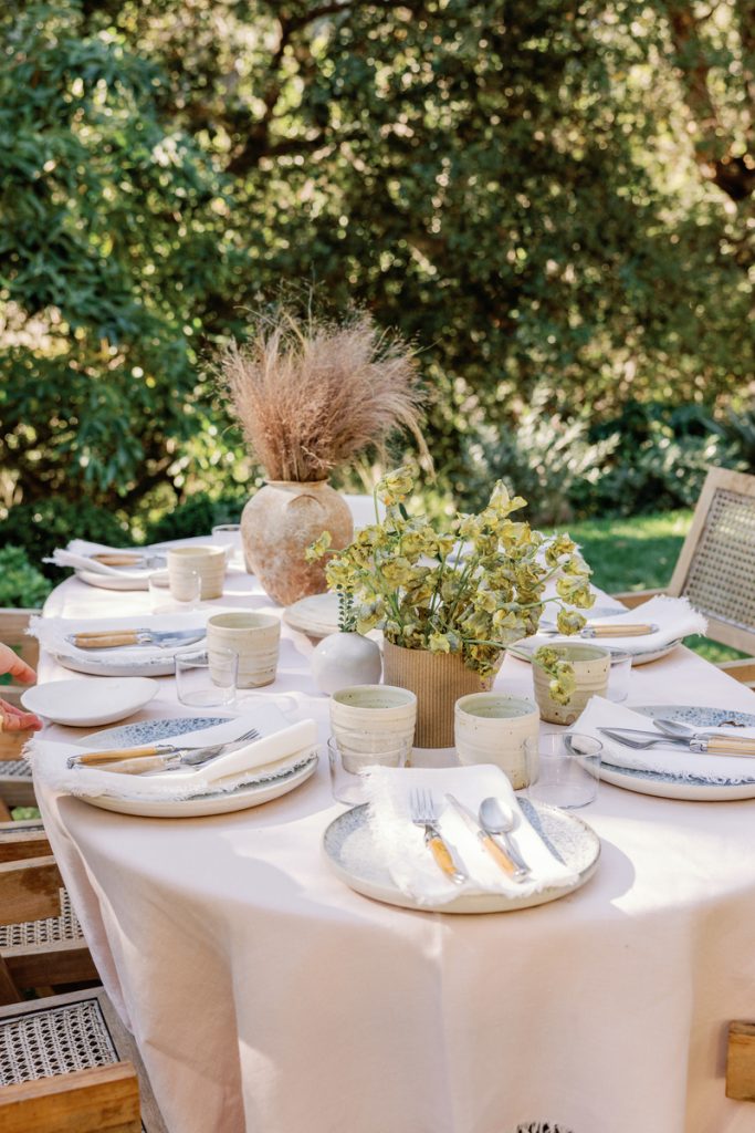 Outdoor dining table set with pink tablecloth, speckled dishware, white napkins, and ceramic/glass glassware.