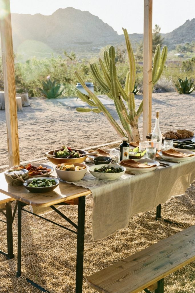 Outdoor desert table setting surrounded by agave plants and cacti with linen tablecloth and various food dishes set on table.