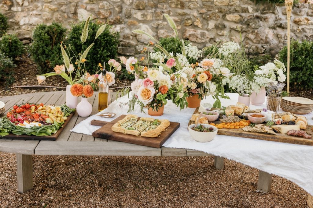 Outdoor table setting with colorful flower bouquets, wooden boards with small bites, and white linen tablecloth.