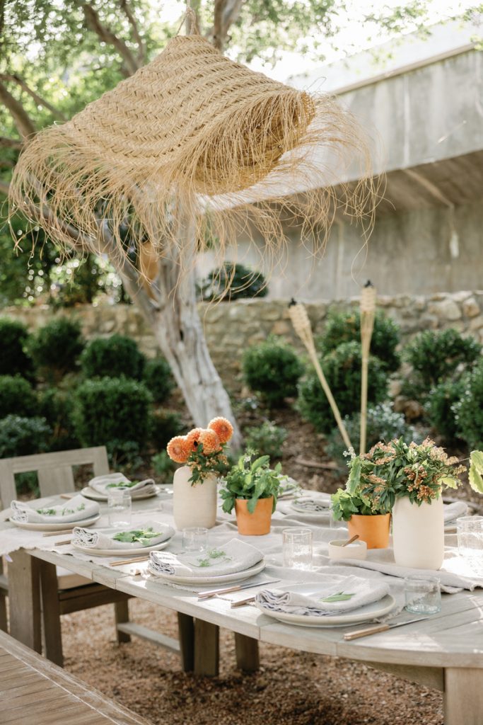 Modern outdoor table setting with white plates, linen tablecloth and napkins, and white vases with dahlias and small plants.