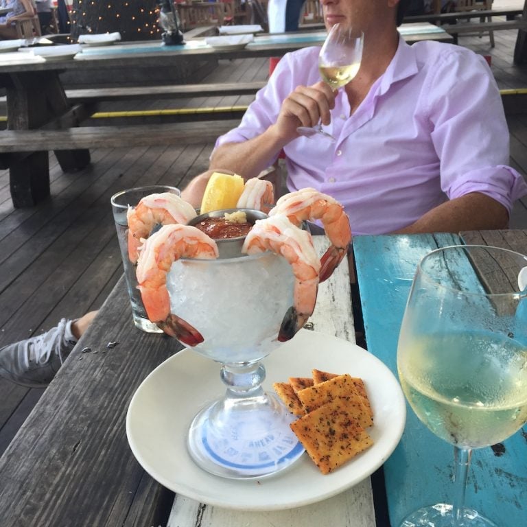 Shrimp cocktail next to glass of white wine on picnic table in front of man wearing purple button down shirt.
