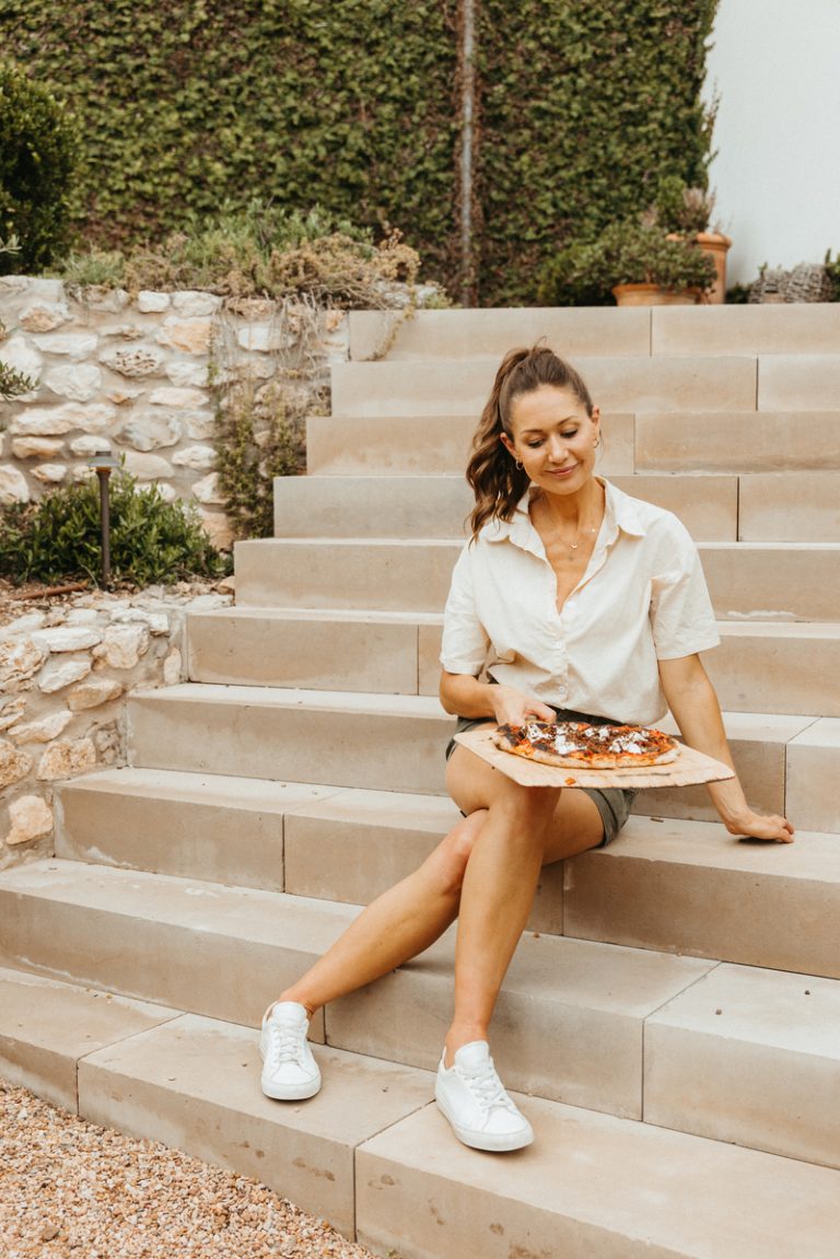 Camille Styles sitting on outdoor steps holding pizza on pizza stone.