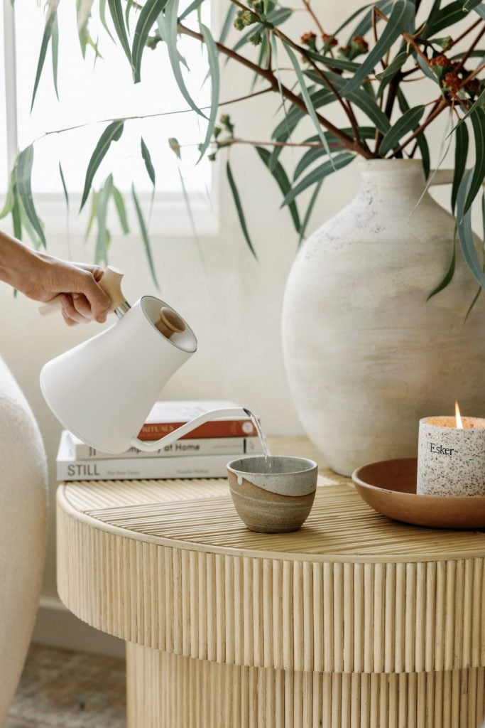 Verter té de una tetera blanca en una taza de té de gres sobre una mesa de café minimalista con jarrón de flores, velas y libros.