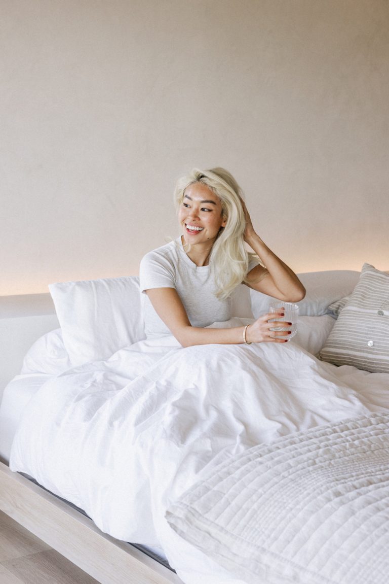 Mulher asiática loira sentada na cama com água potável de lençóis brancos.