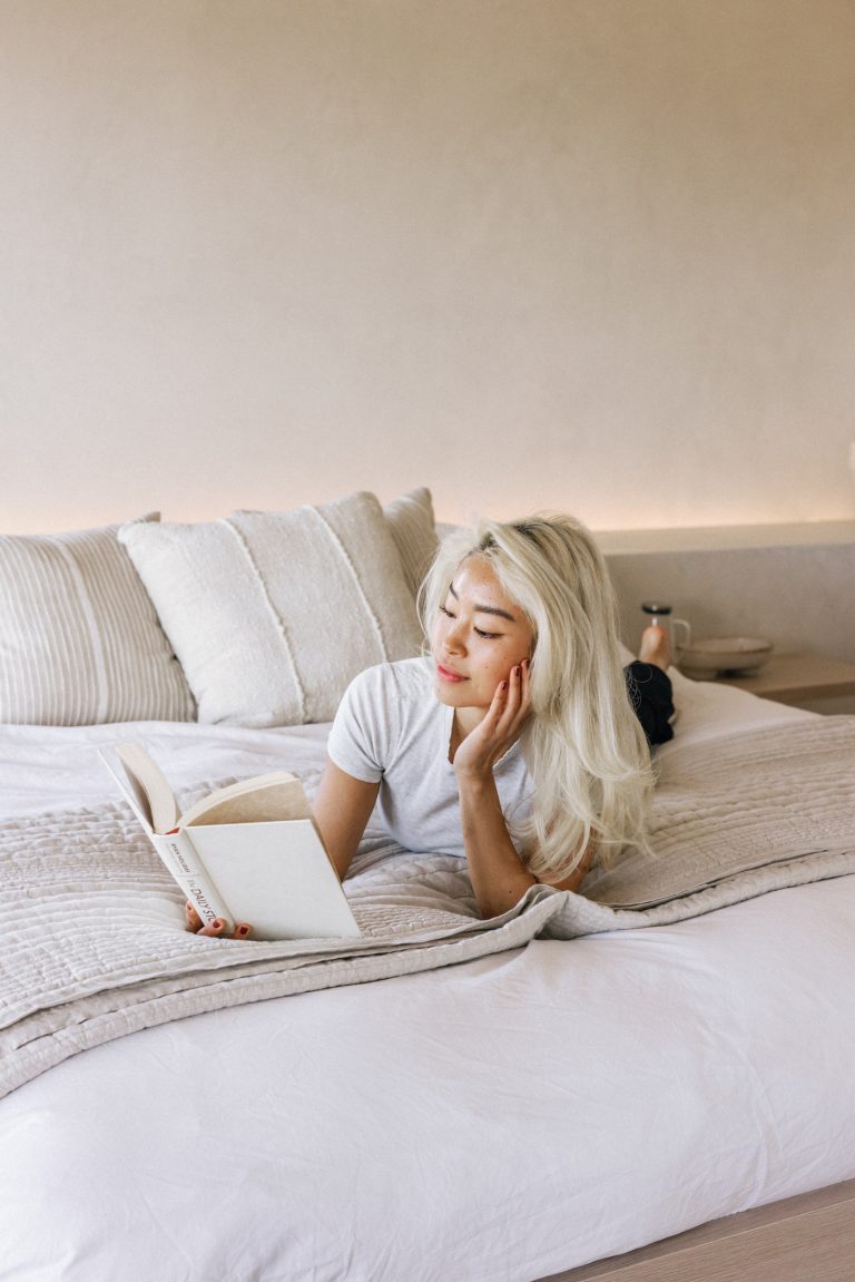 Blonde Asian woman reading book on bed with white sheets.