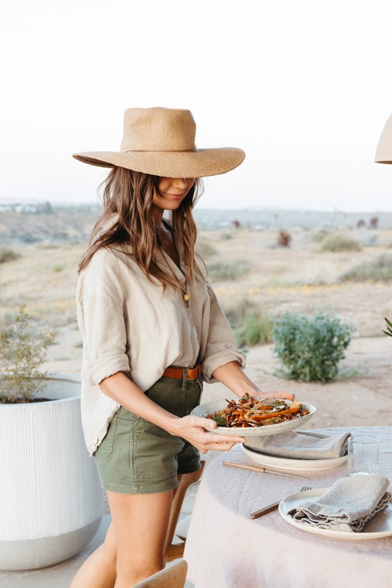 Brunette woman wearing sun hat serving a plate of roasted carrots outside.