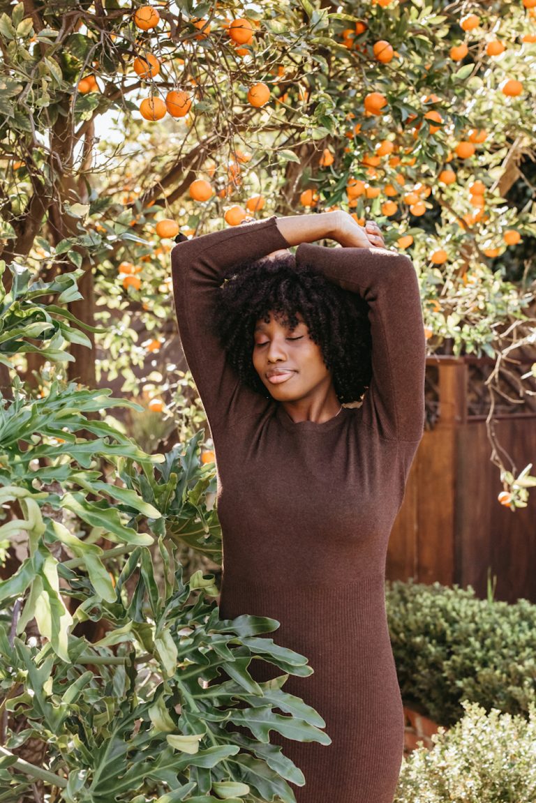 Black woman wearing long-sleeved brown sweater dress standing outside in front of orange tree.