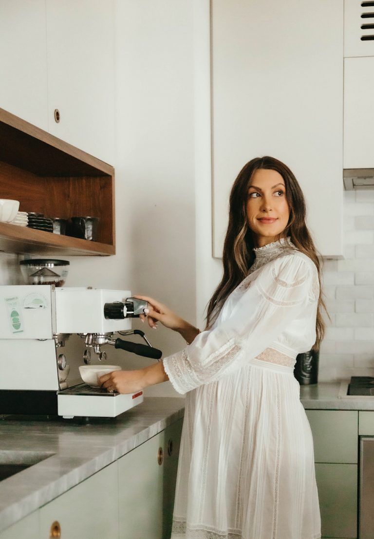 Brunette woman in long white cottagecoa dress using espresso machine in kitchen.
