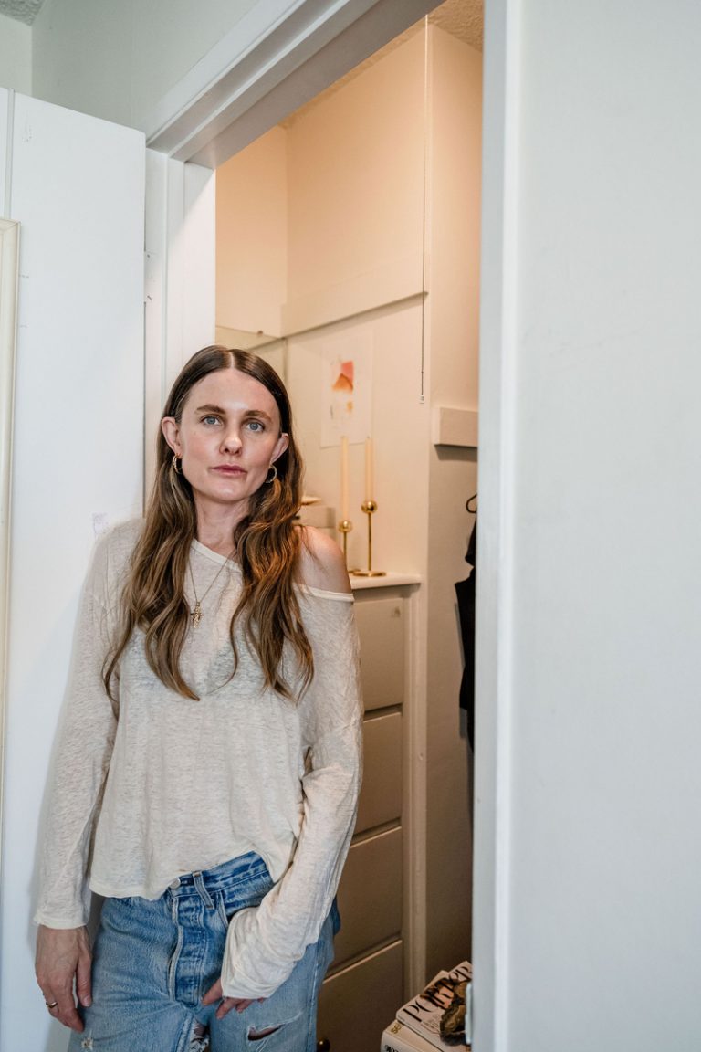 Sacha Strebe wearing slouchy white sweater and jeans leaning against white wall outside of home closet.