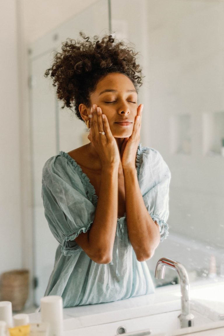 Black woman applying skincare to face in bathroom mirror.