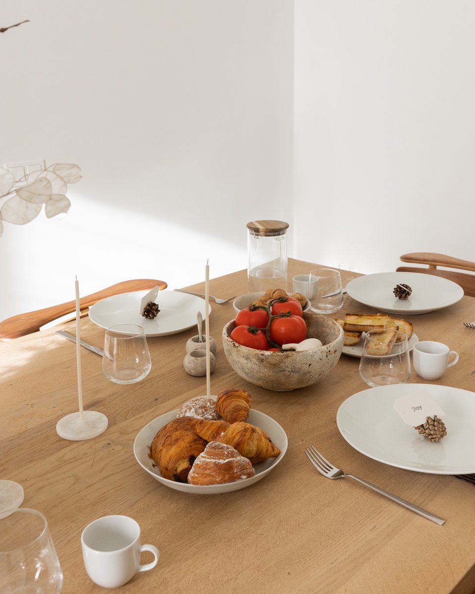 Simple Scandinavian table setting with white plates, pinecone place cards, pastries, white candlesticks, and bowl of fruit.