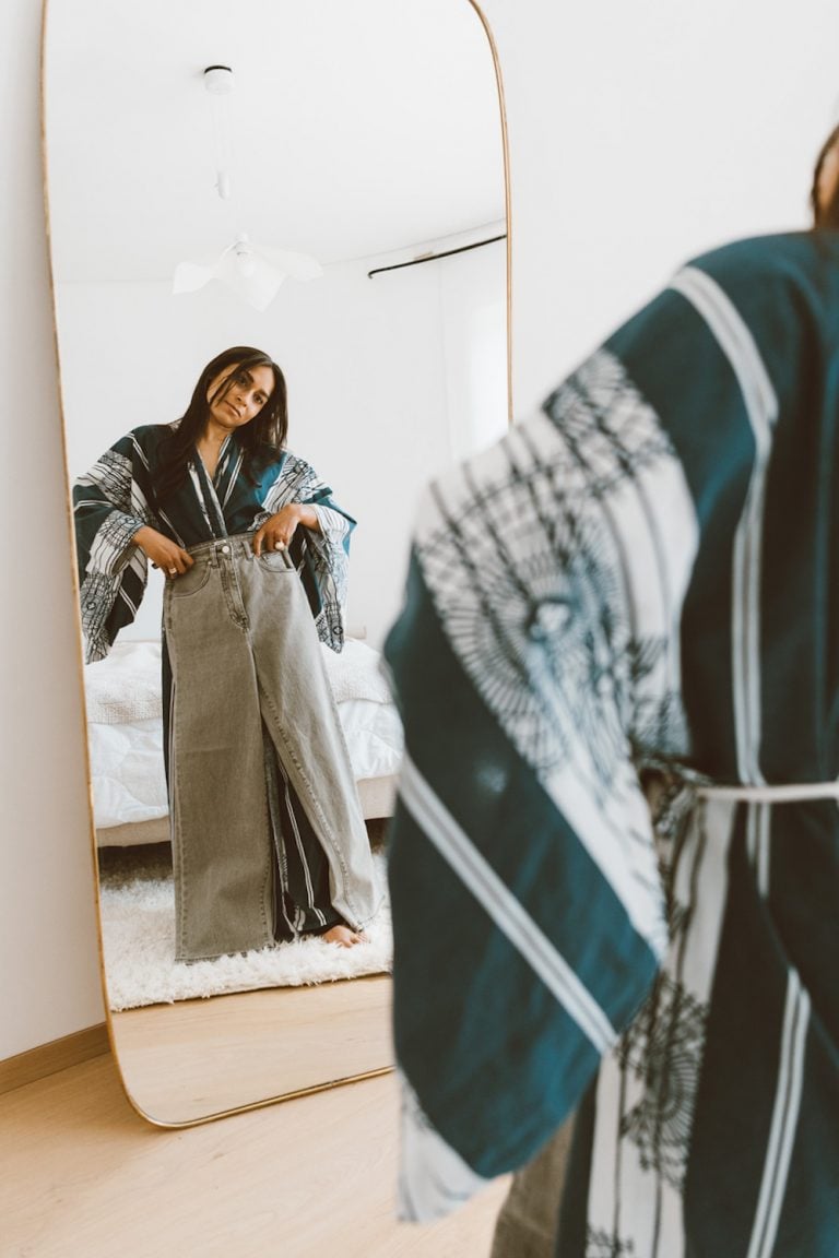 Brunette woman wearing robe holding jeans up to body in mirror.