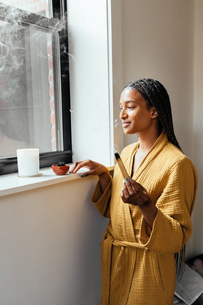 A black woman with long braids in a yellow robe illuminating Palo Santo.