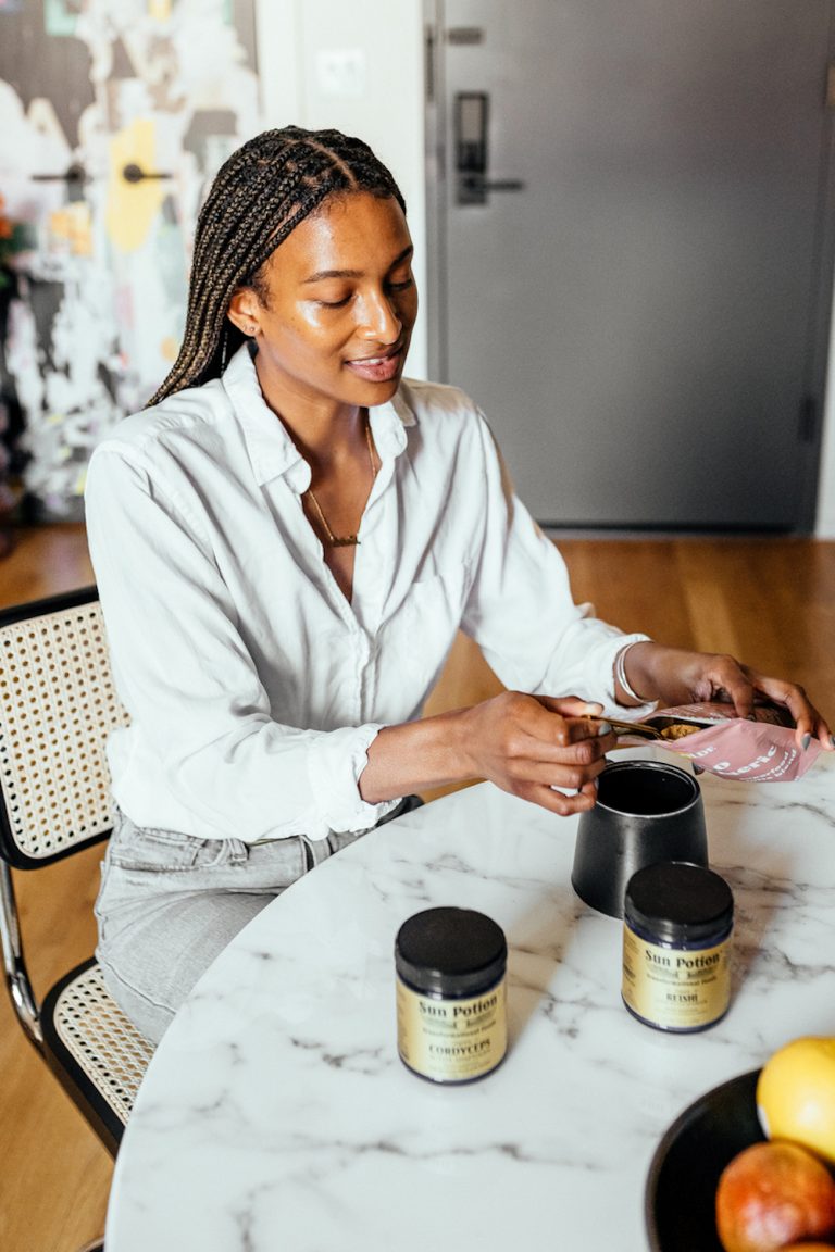 Black woman wearing white button-down shirt and jeans measuring out health supplements at marble kitchen table.