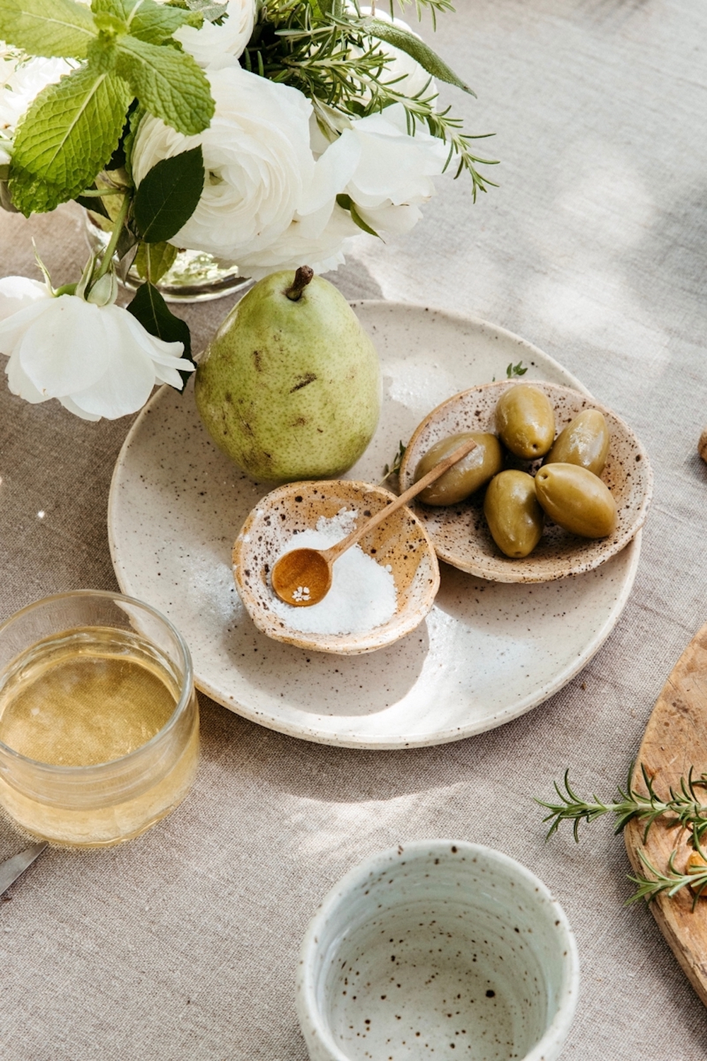 Stoneware serving dishes filled wish flaky salt and olives on stoneware plate with pear next to glass of wine and bouquet of white flowers.