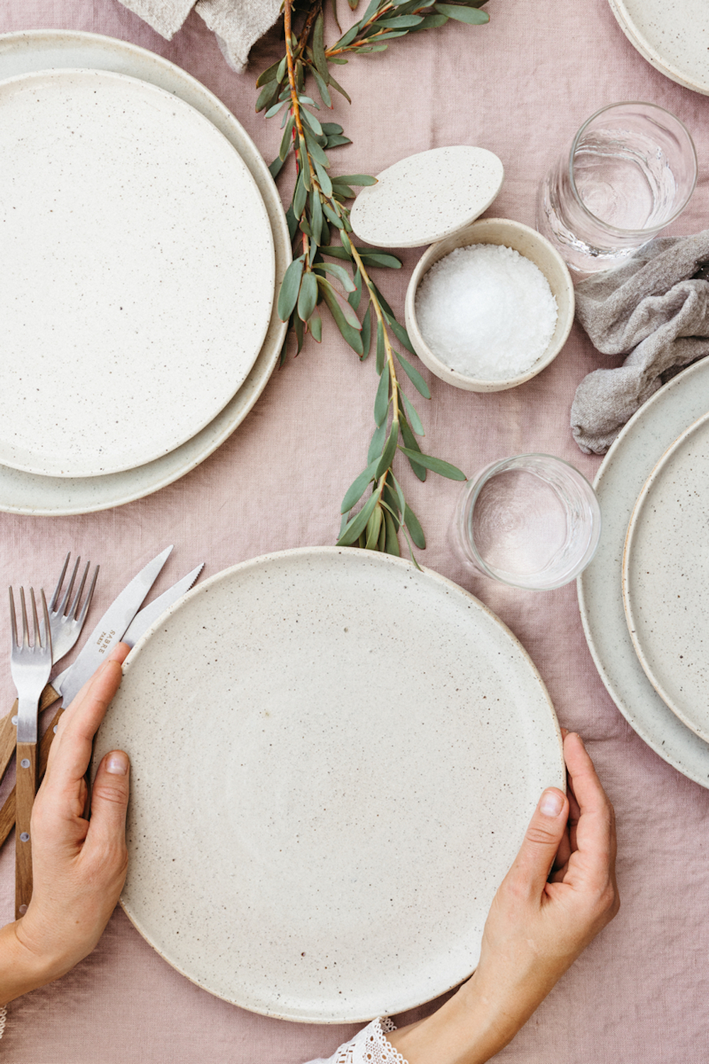 Hands setting down speckled stoneware plate on pink tablecloth with other neutral plates, salt cellar, glassware, and flatware.