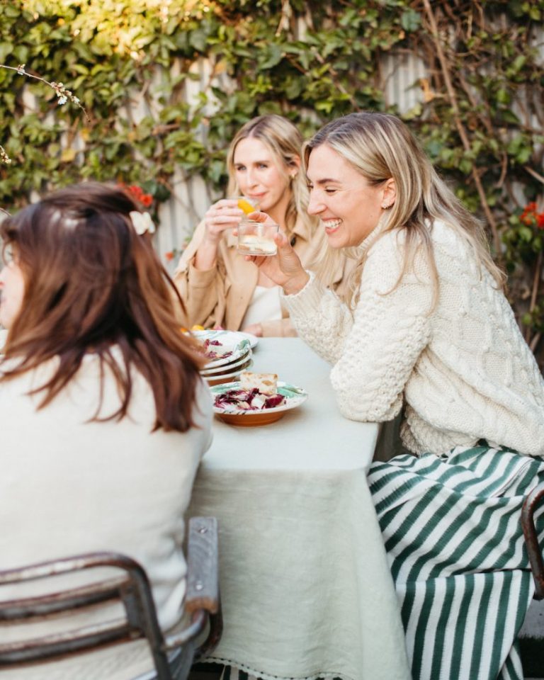 Three women drinking cocktails laughing at outdoor dinner table.