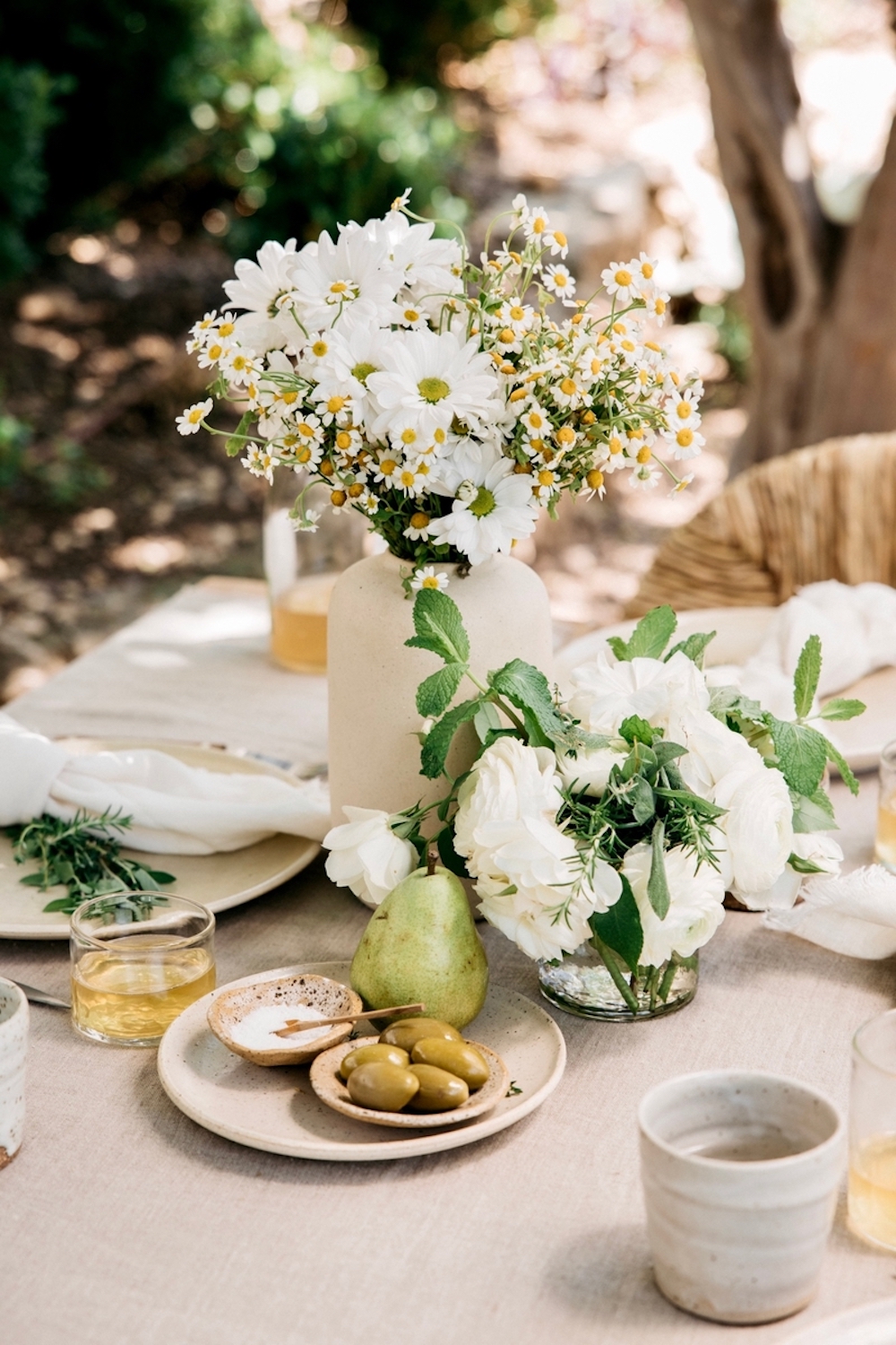Outdoor spring table setting with pink linen tablecloth, centerpiece with small bowls of salt, olives, and a green pear, a small white flower centerpiece, and a speckled stoneware vase of daisies and white flowers.