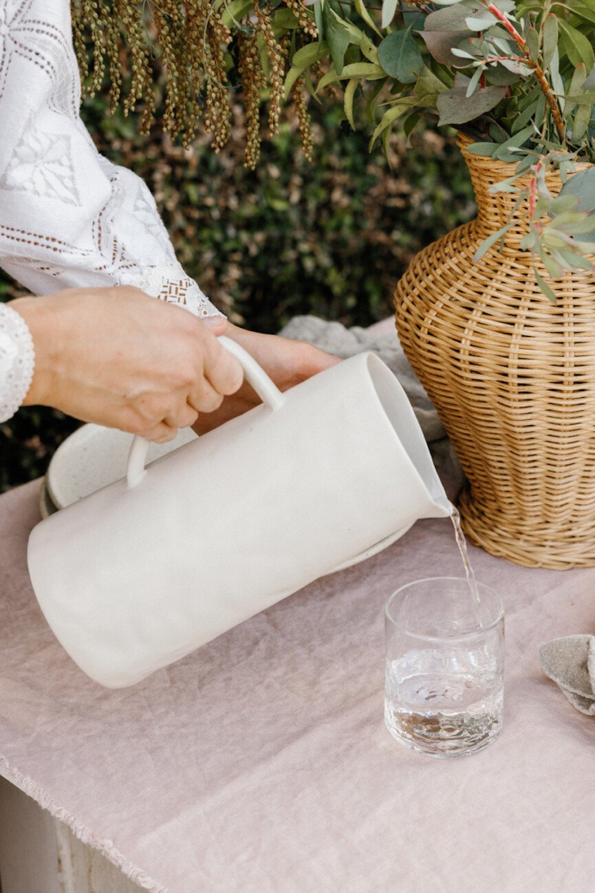 Woman pouring stoneware carafe of water into clear water glass on pink linen tablecloth.