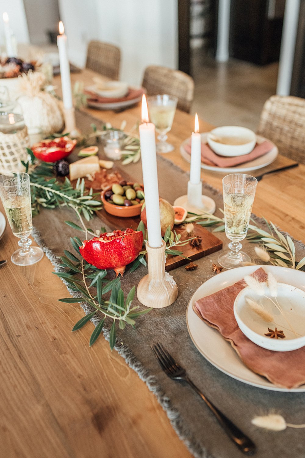 Ideas para poner la mesa del Día de Acción de Gracias con un camino de mesa gris oscuro, velas cónicas blancas, cristalería estriada, ramitas de olivo y vajilla neutra.