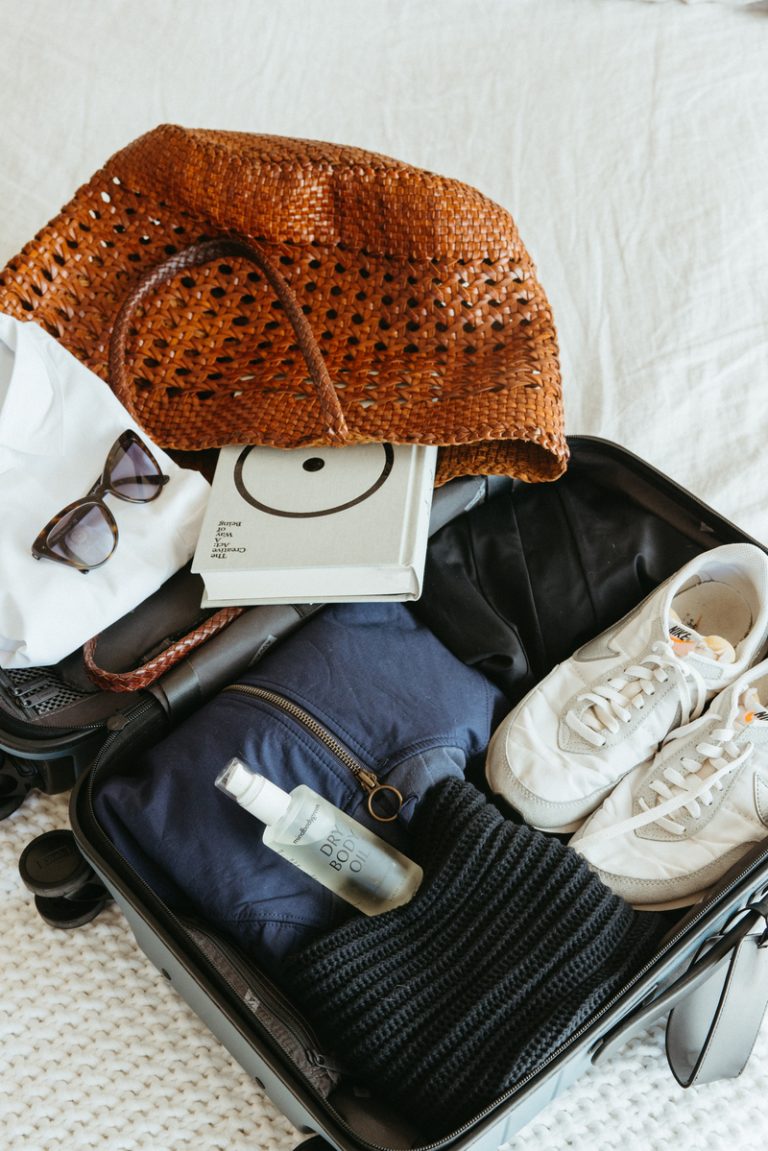 چمدان روی تخت بسته بندی شده با لباس، کتاب، کفش ورزشی، کیف پول، و عینک آفتابی باز است.