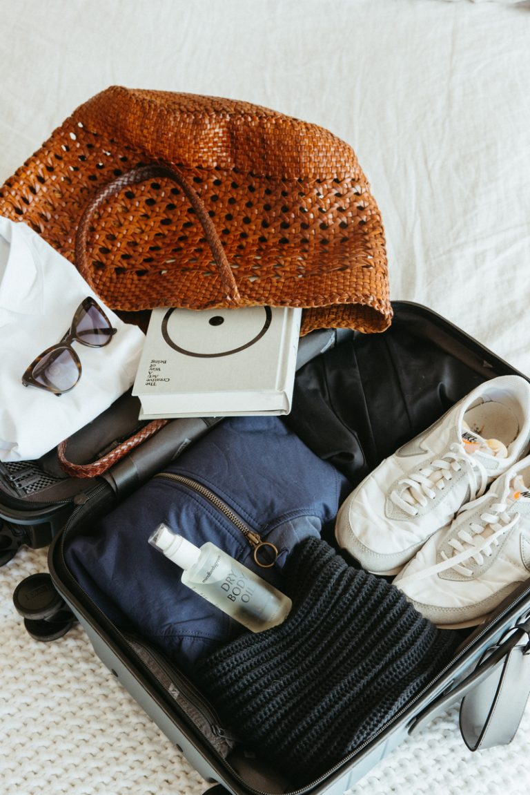 Suitcase open on bed packed with clothing, book, sneakers, purse, and sunglasses.