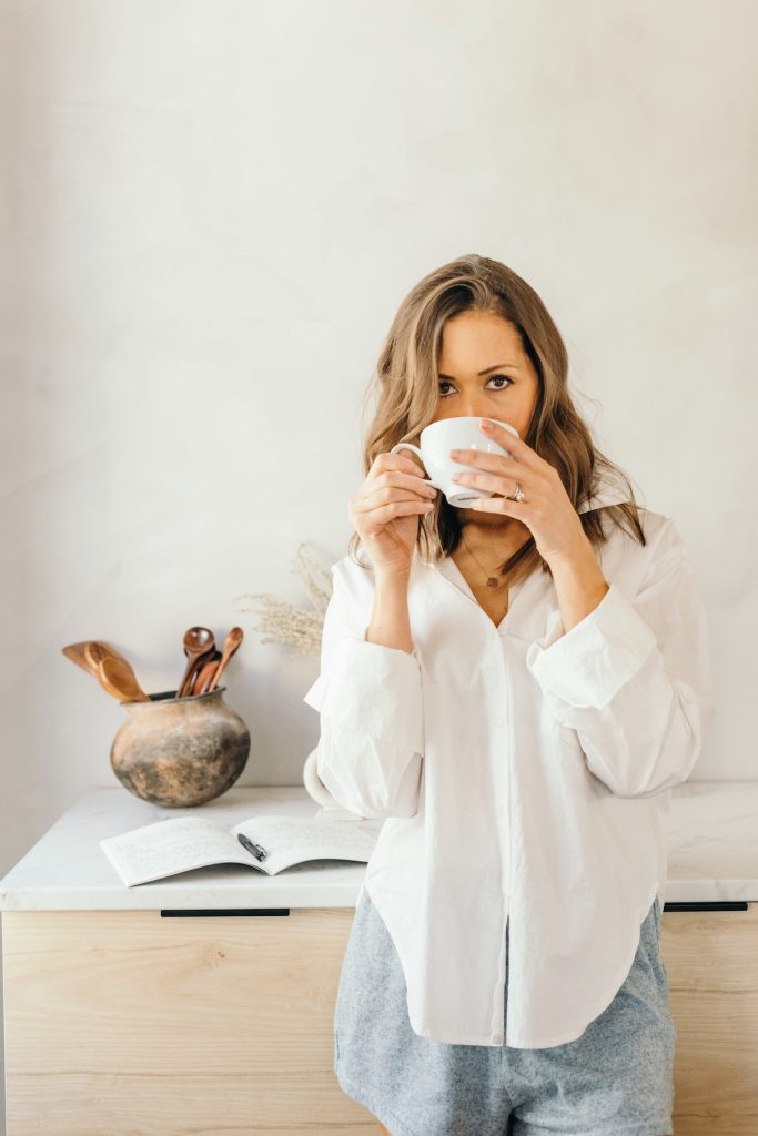 Camille Styles wearing white button-down shirt drinking tea at kitchen counter.