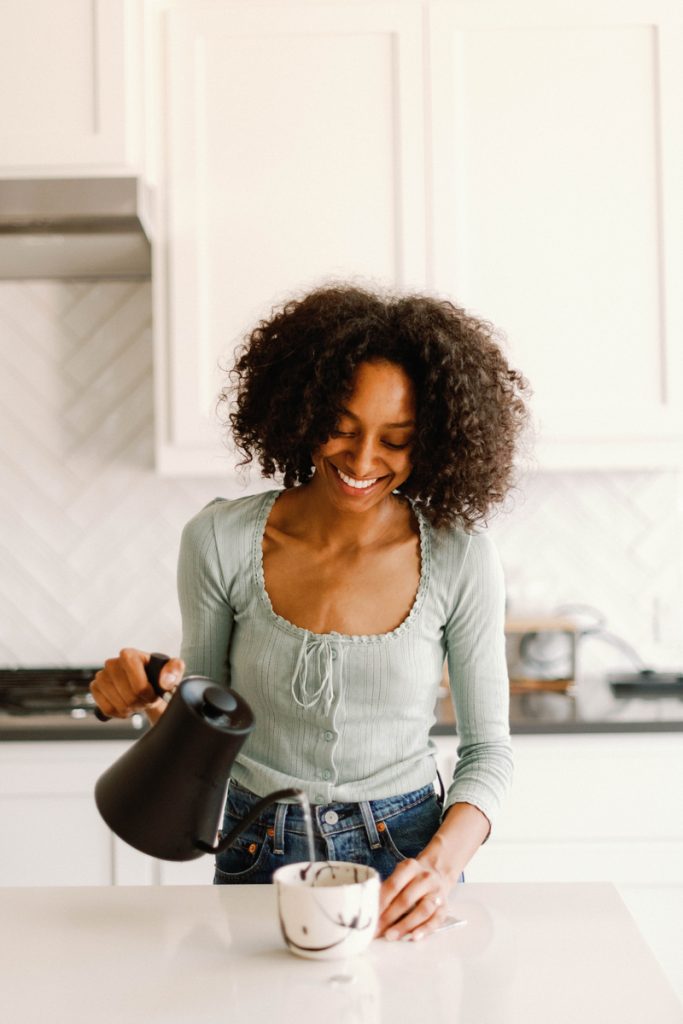 Black woman wearing blue long-sleeved shirt pouring hot water from black kettle into mug of tea on white kitchen counter.