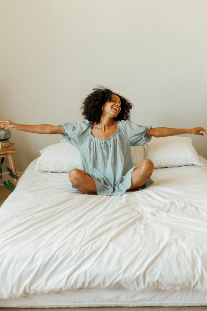 Mujer negra con camisón azul sonriendo y extendiendo los brazos mientras se acuesta en la cama con sábanas blancas.