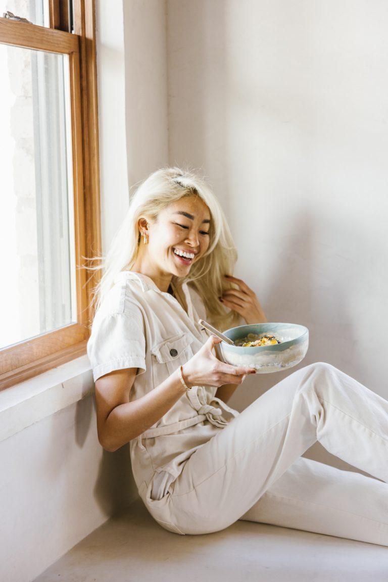 Blonde woman smiling wearing white jumpsuit holding bowl of oatmeal.
