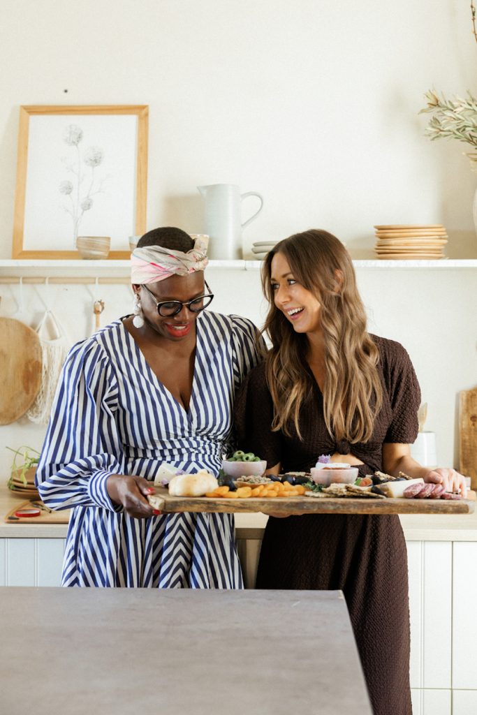 Camille Styles and Ajiri Aki holding cheeseboard in kitchen.