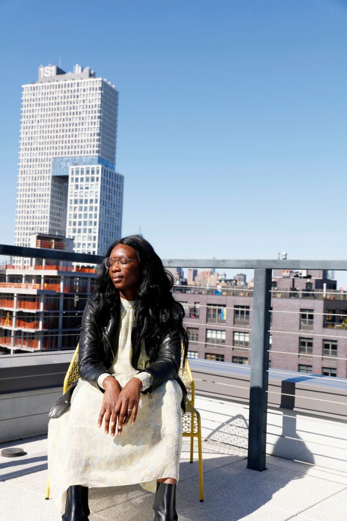 Black woman wearing leather jacket and dress sitting on city rooftop.