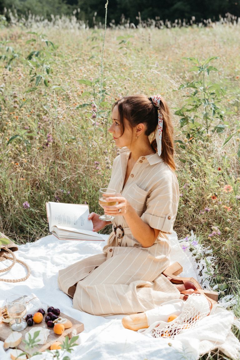 Brunette woman drinking glass of wine and reading in field on picnic blanket.