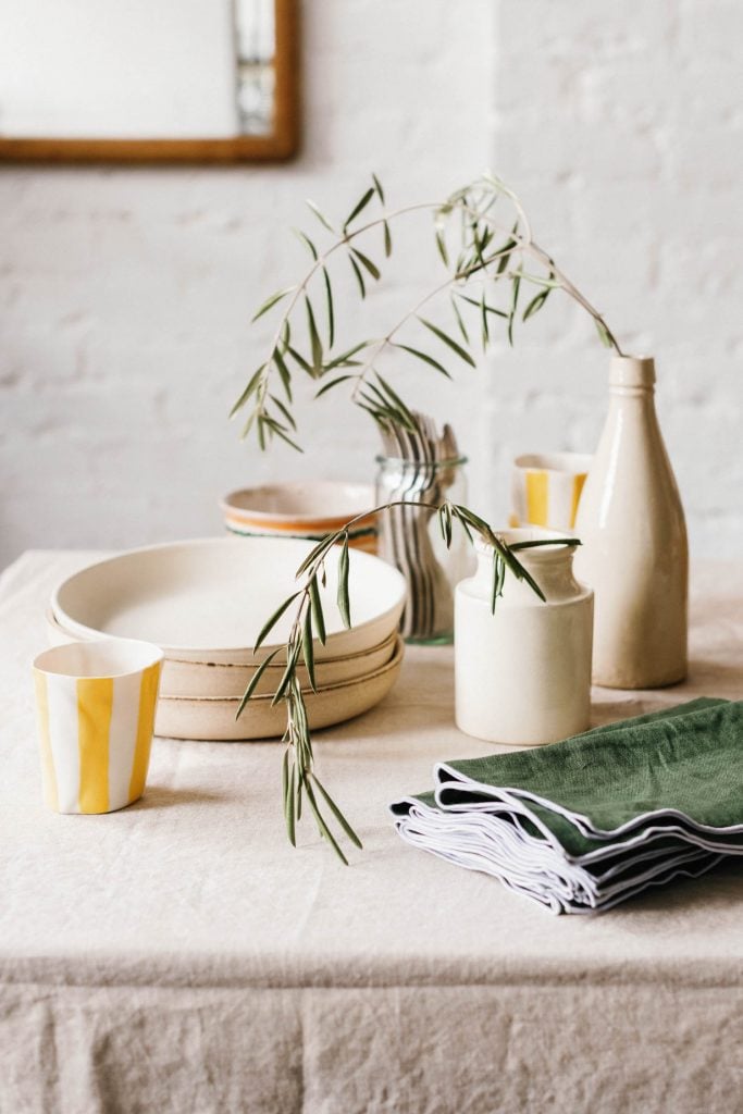 Mesa minimalista de linho com pratos de cerâmica, xícaras e vasos cheios de ramos de oliveira.