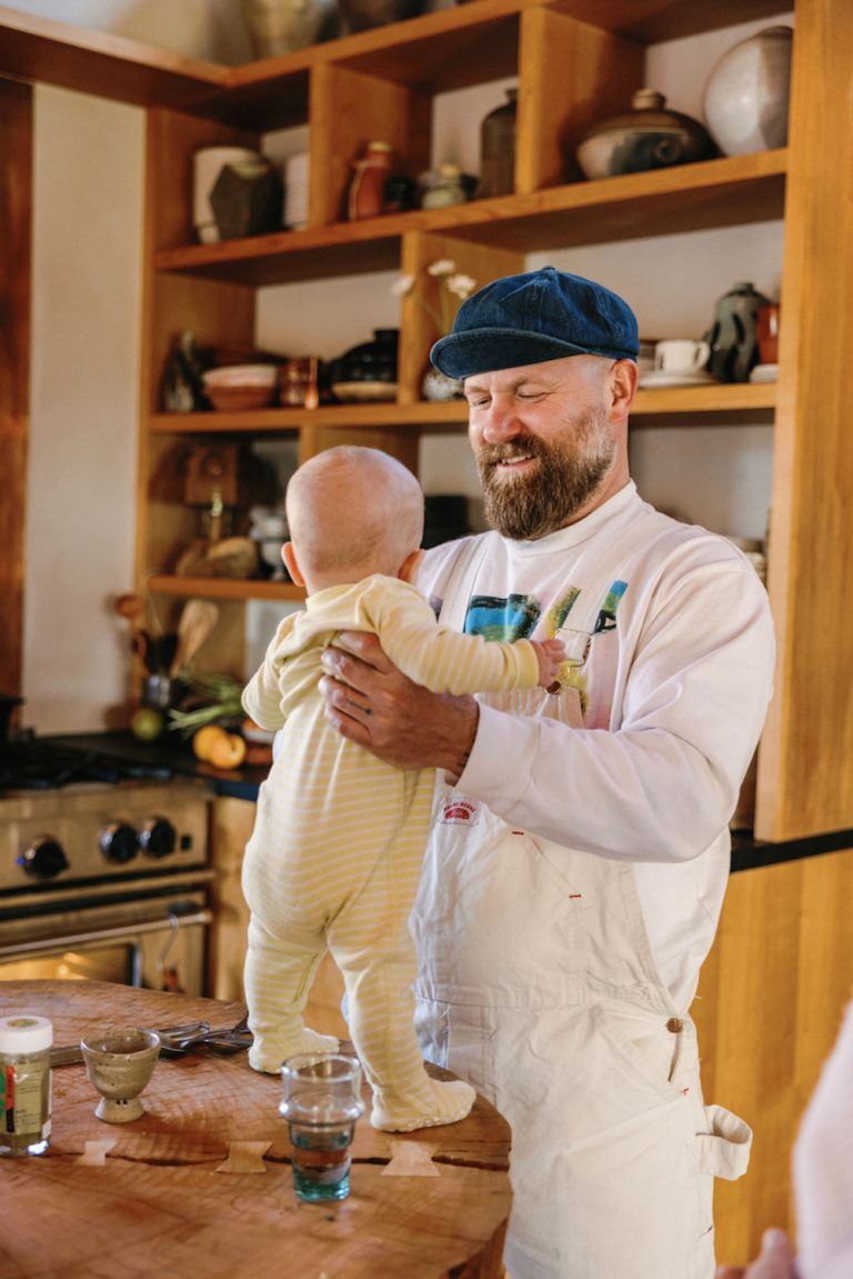 Man wearing white overalls and hat holding baby wearing yellow onesie.