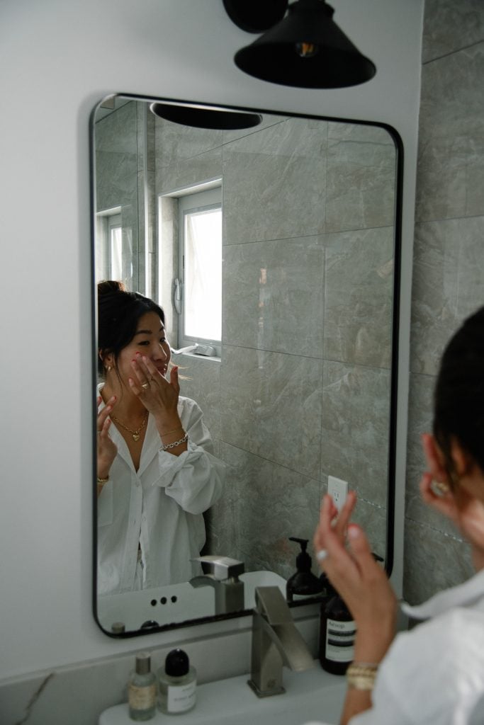 Asian woman applying skincare product in bathroom mirror.