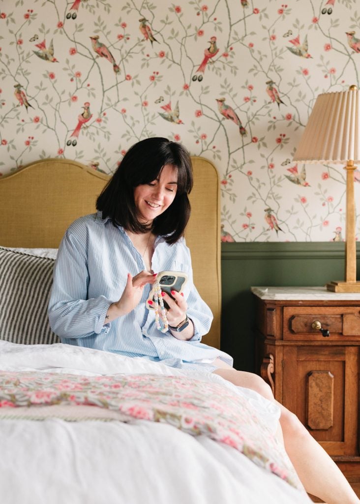 Brunette woman wearing white striped button-down shirt using phone while sitting on bed.