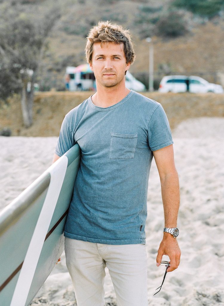 A man in a blue shirt and pants standing on the beach with a surfboard.