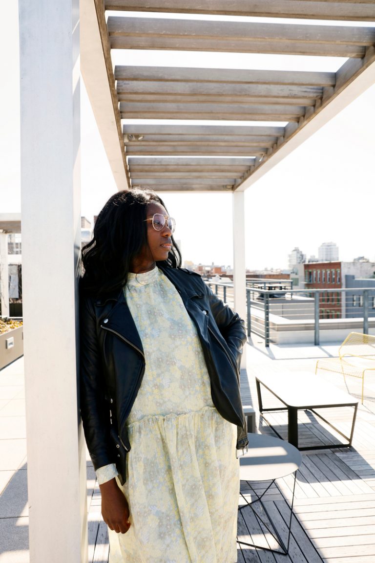 Black woman wearing yellow floral dress and leather jacket standing outside.